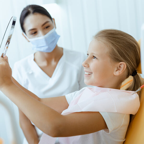 Young girl smiling at her reflection in a handheld mirror during a dental appointment.