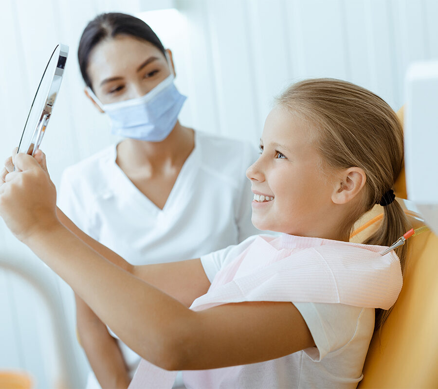 Young girl smiling at her reflection in a handheld mirror during a dental appointment.